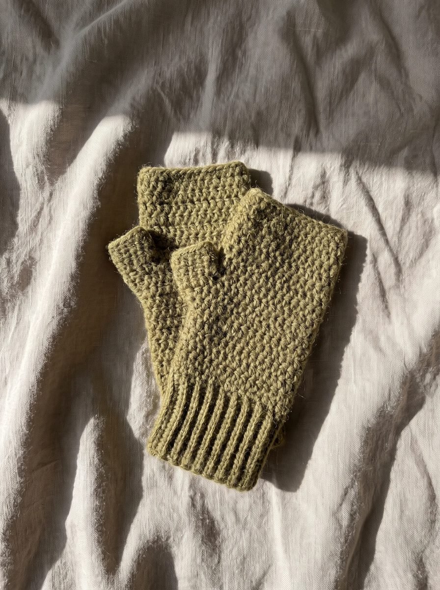 Pair of olive green crochet fingerless mitts with ribbed cuffs lying on rumpled white linen in a stripe of sunlight