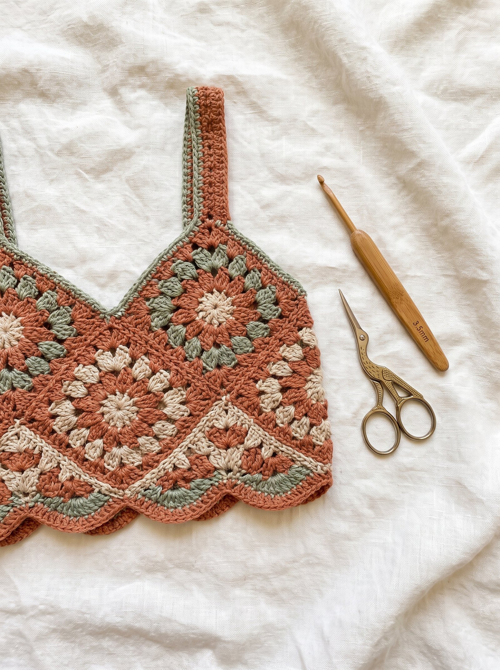 A bohemian crochet granny square bralette in terracotta, sage green, and oatmeal laid flat on an ash wood workbench beside a bamboo hook