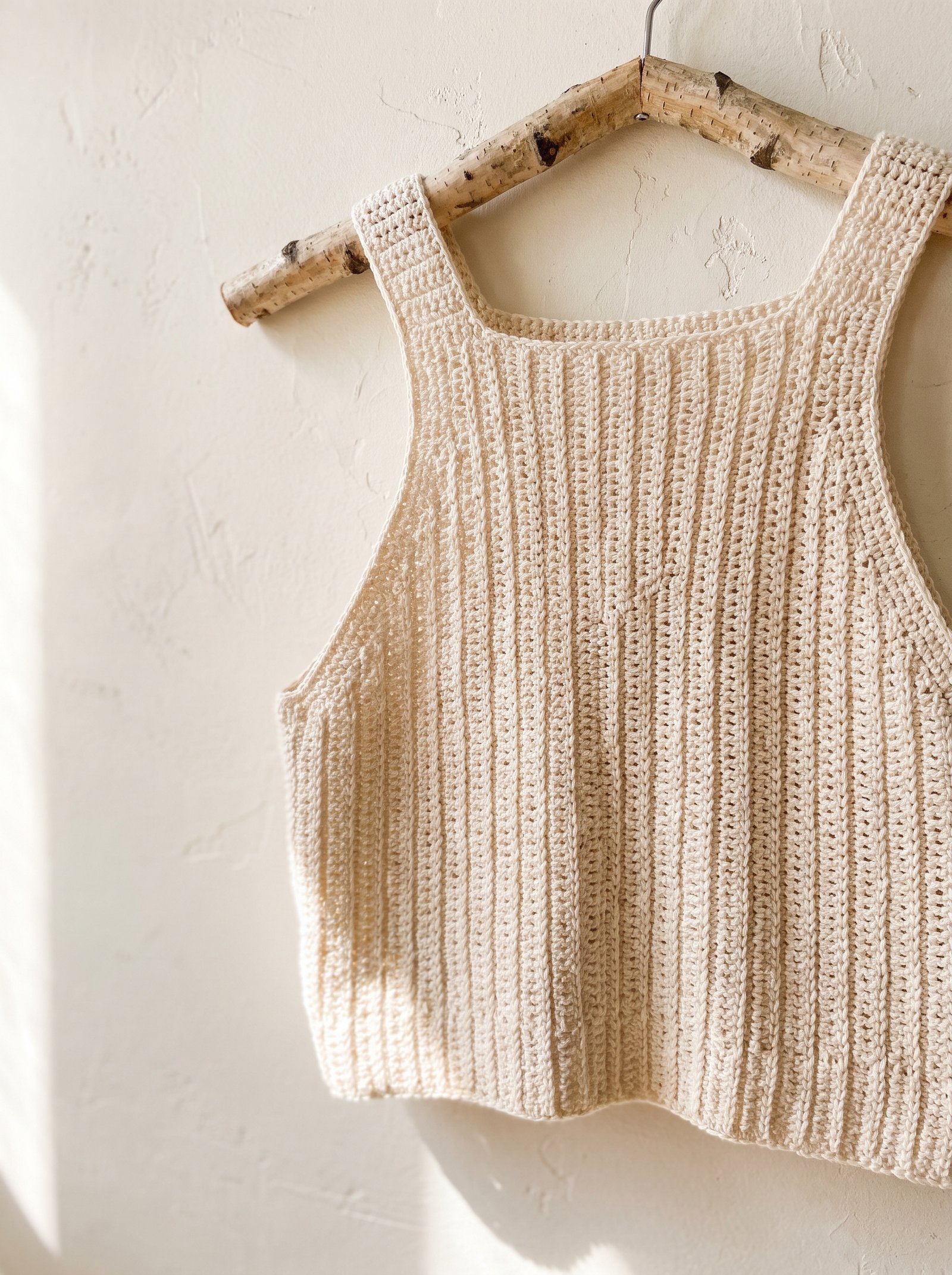 A modern cream crochet crop camisole with clean vertical ribbing and square neckline hanging from a birch branch hanger against a textured wall