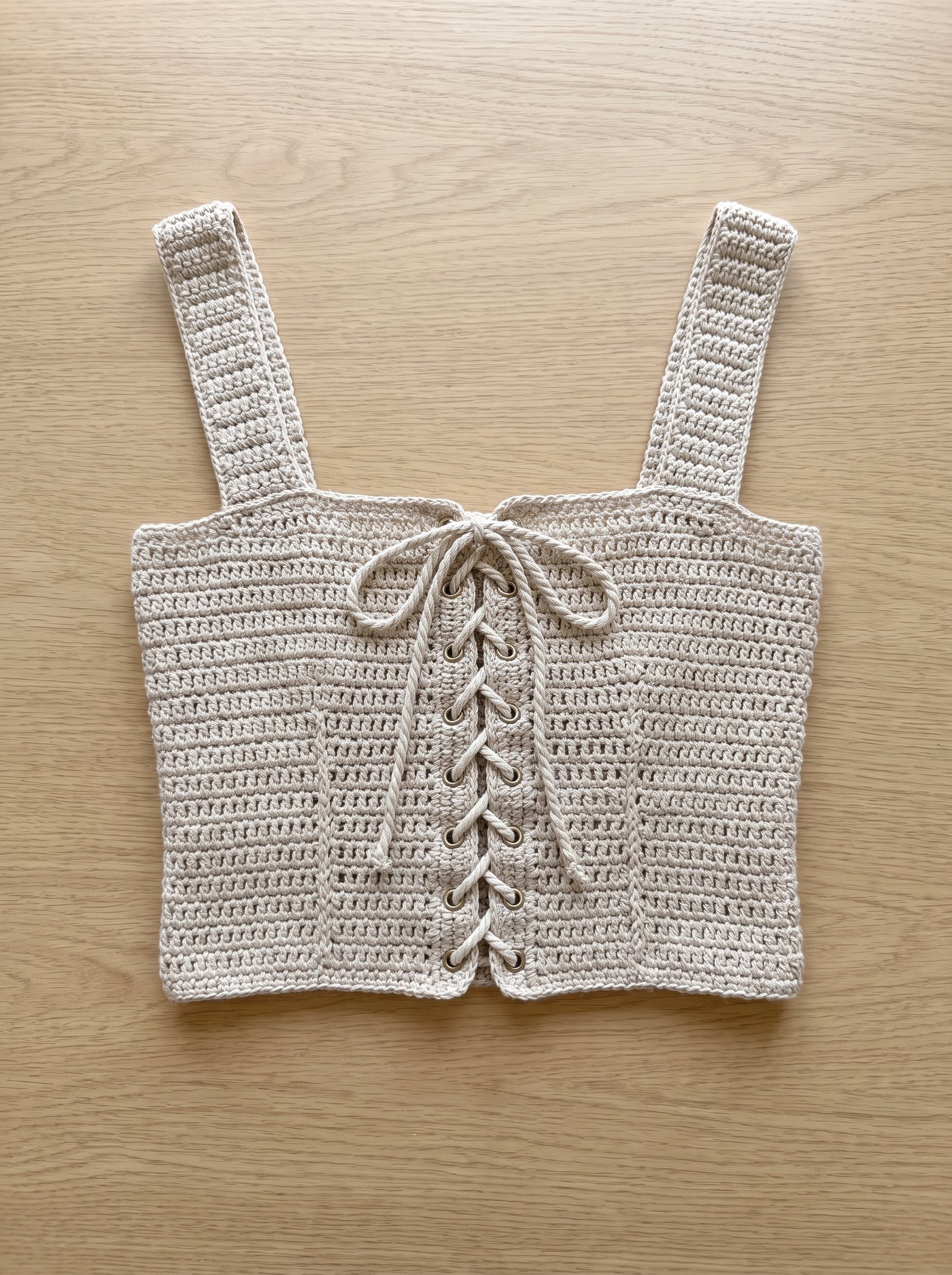 A structured sand-toned crochet corset crop top with eyelet lace-up back laid flat across a weathered oak studio table with wooden hook