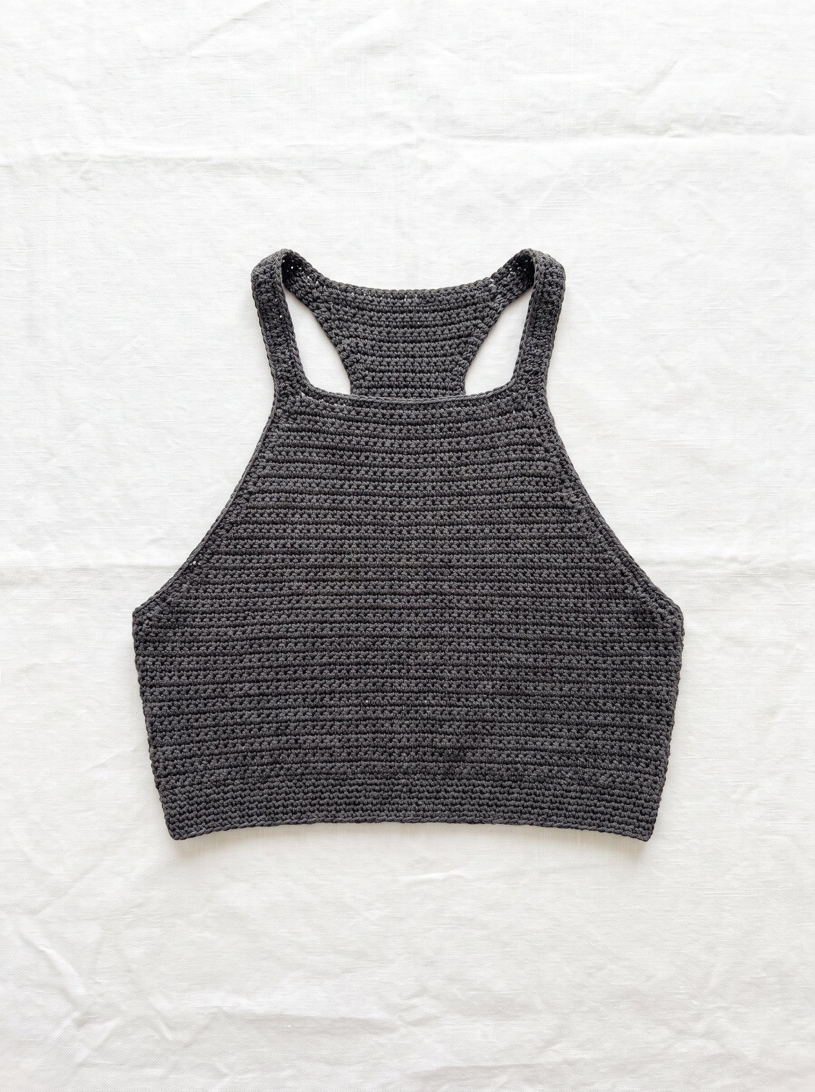 A sleek dark charcoal athletic crochet crop top with high sporty racerback neckline laid flat across textured white linen fabric