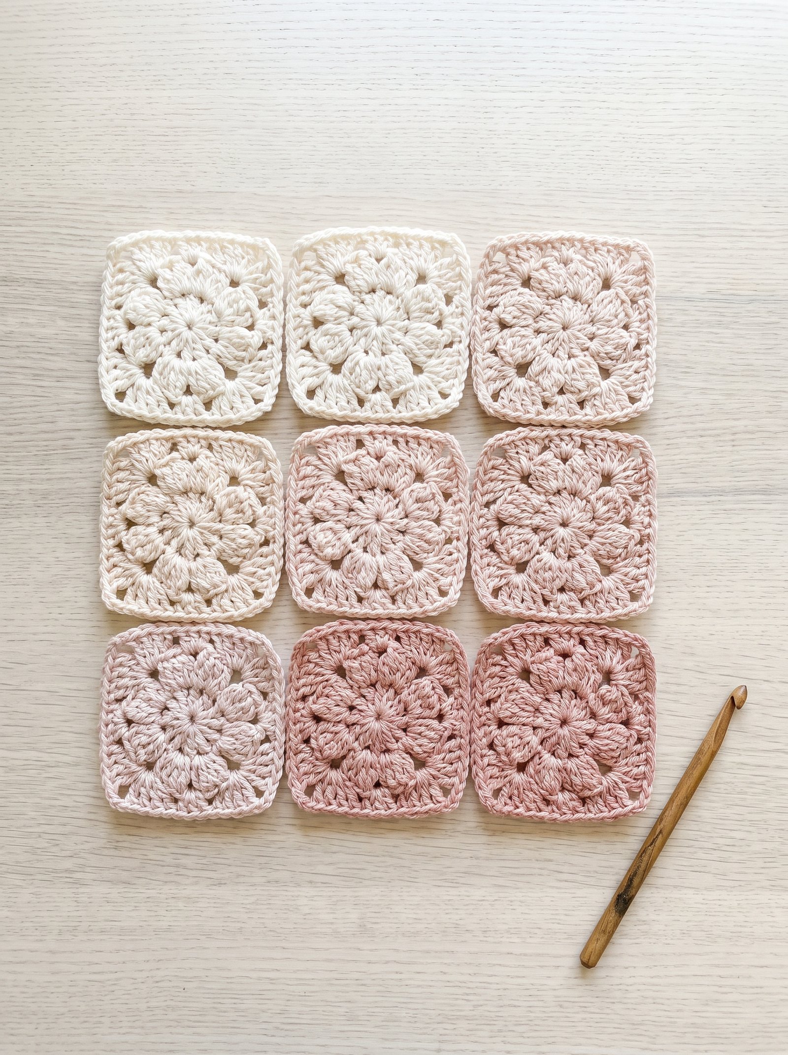 Nine monochromatic floral crochet squares arranged in a gradient ombre from charcoal to silver on a pale birch table with an aluminum hook