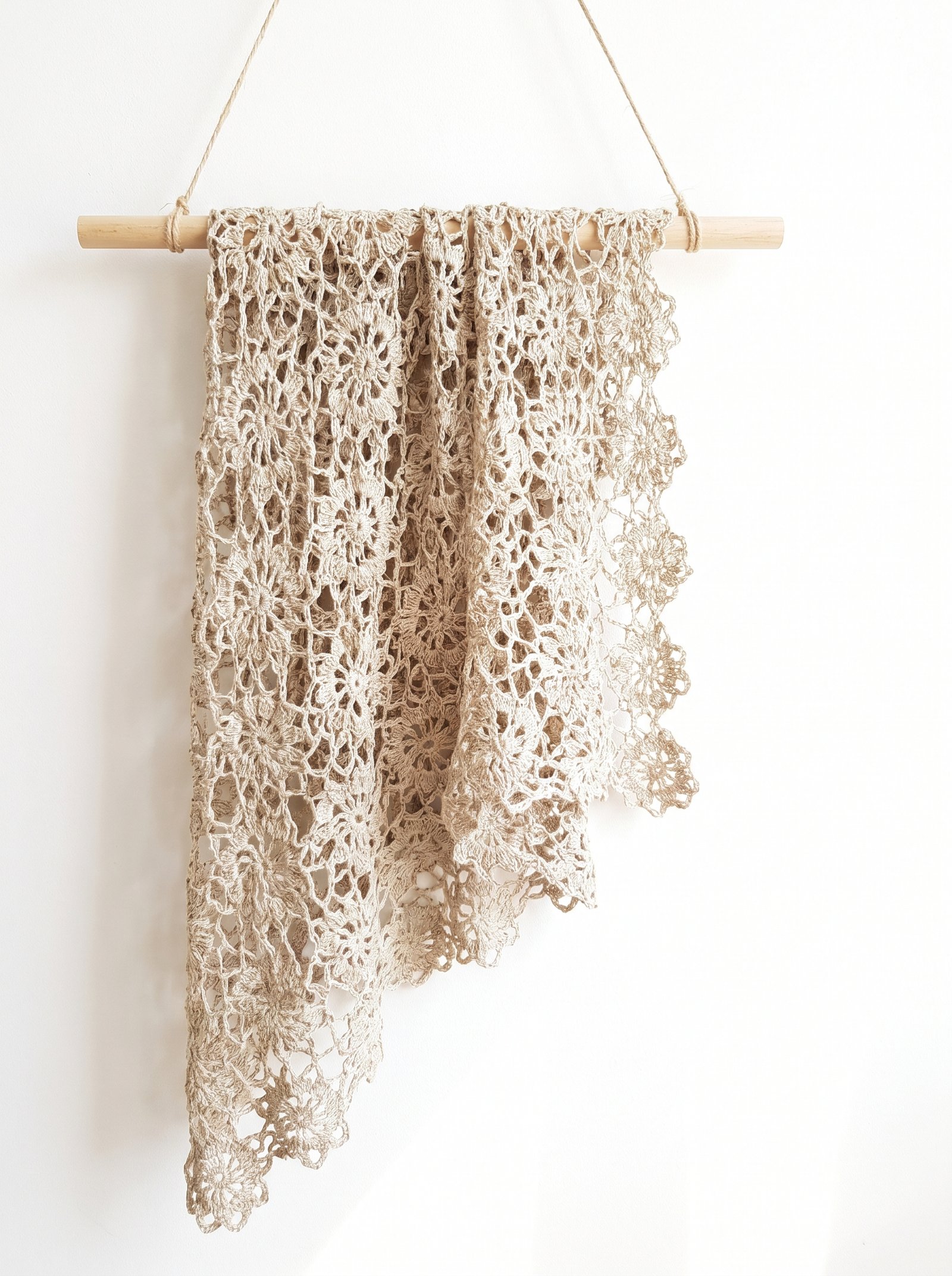A lightweight openwork linen lace floral crochet throw draped gracefully over a natural wooden wall dowel hung with simple jute cord