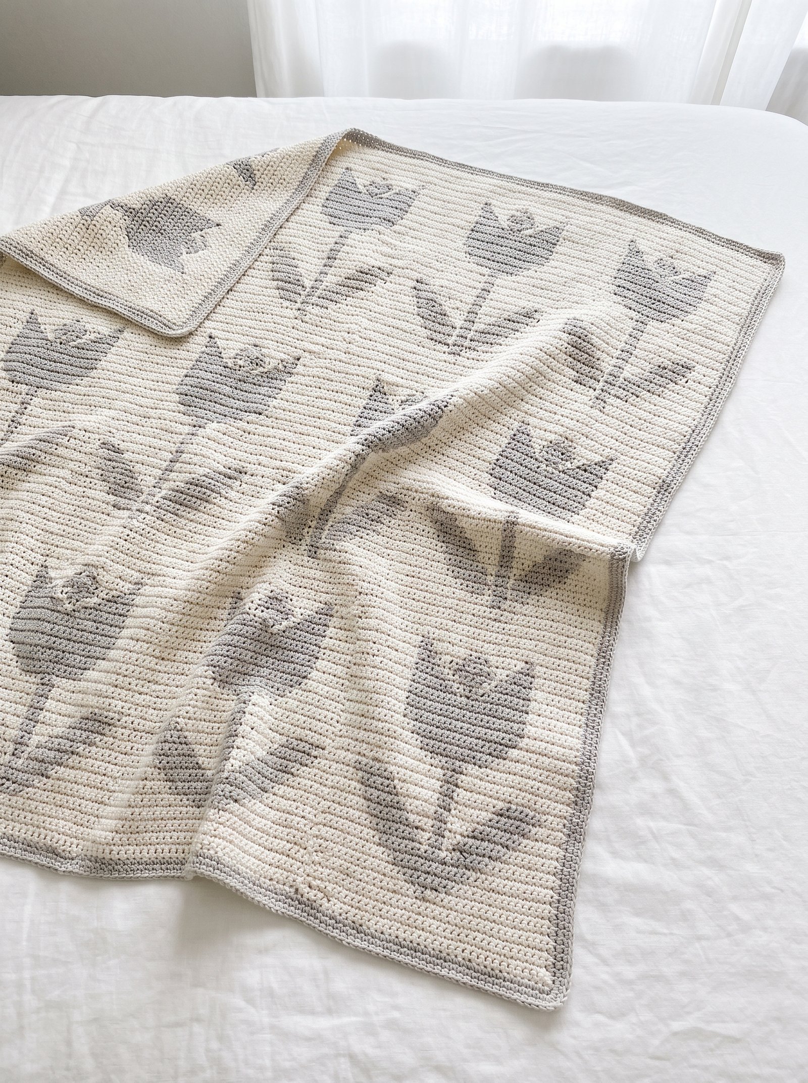 A modern two-tone tapestry crochet tulip blanket in cream and warm taupe laid flat across clean white bed linen in soft daylight
