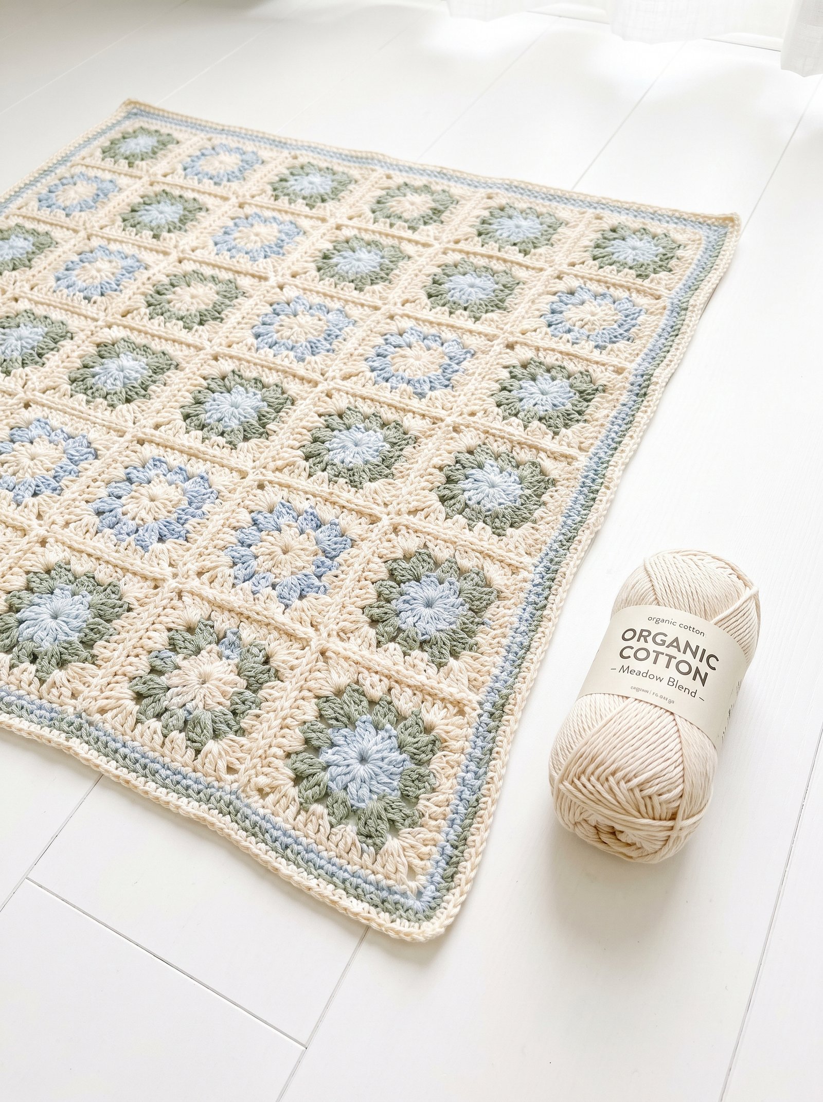 A delicate pastel meadow crochet flower blanket laid out on a pale pine floor beside a fresh skein of organic unbleached cotton yarn