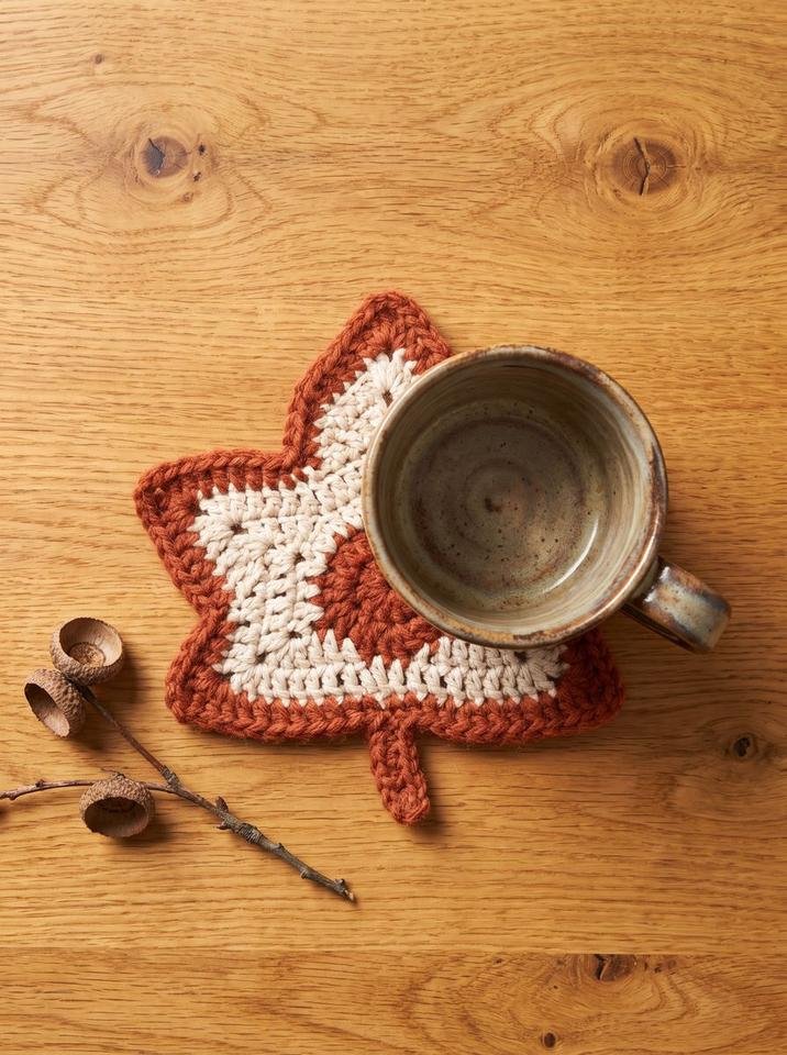 Two-tone crochet maple leaf mug rug with cream center and terracotta edging holding a handmade ceramic cup on an oak table