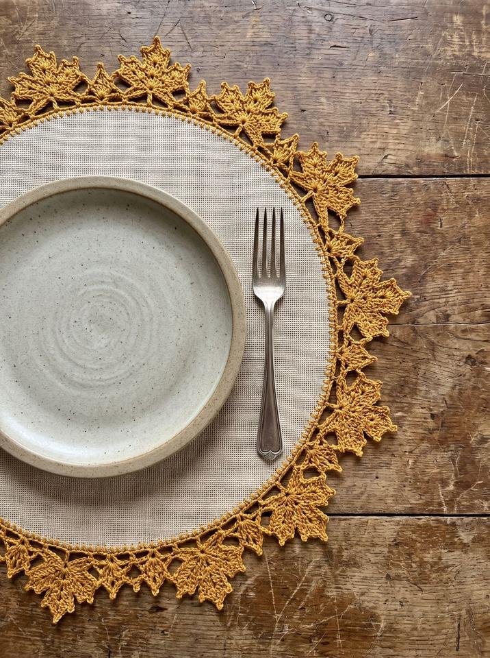 Circular placemat with woven fabric center and scalloped golden crochet maple leaf edging beside ceramic tableware
