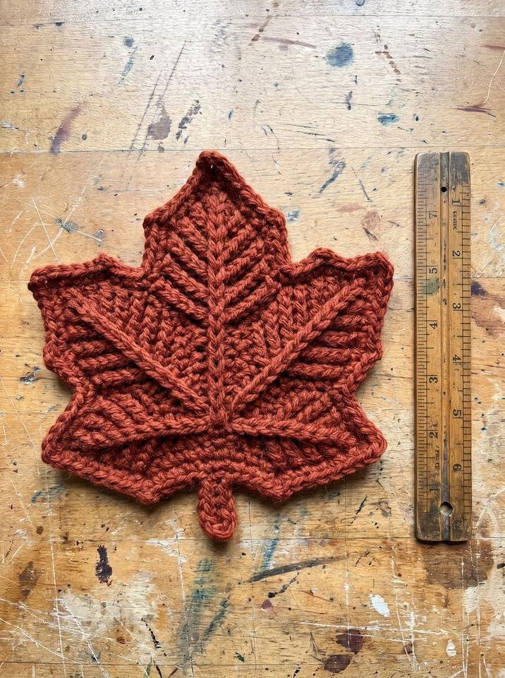 Large thick crochet maple leaf hot pad with raised front post veins in rust cotton measured next to a vintage wooden ruler on a workbench