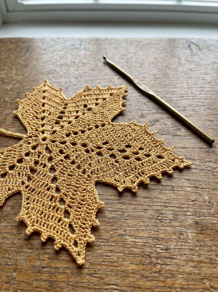 Delicate lace crochet maple leaf doily in golden thread with fine picots next to a vintage brass hook on an oak table