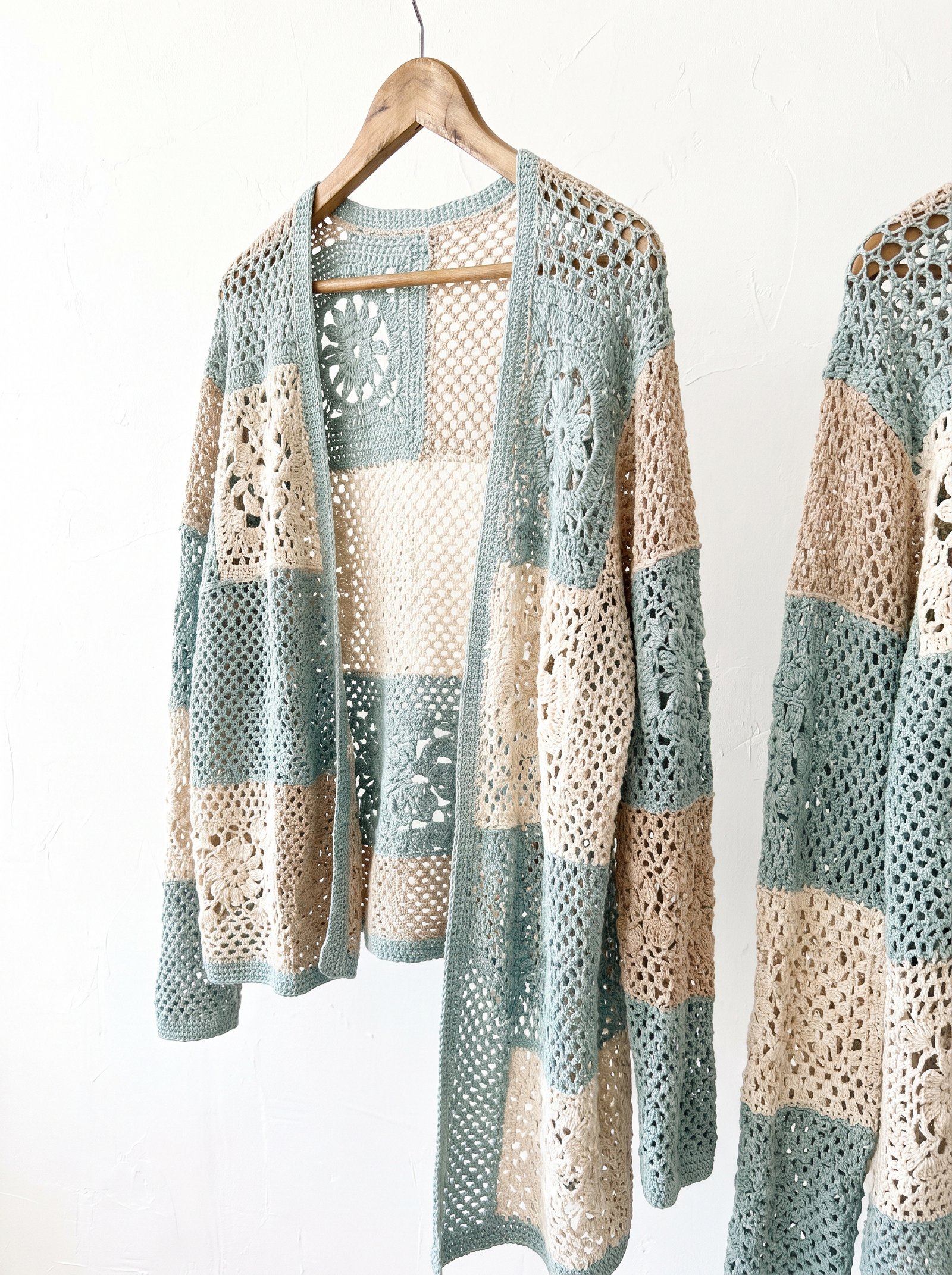 A cropped monochrome crochet patchwork cardigan with cream and charcoal checkerboard squares laid flat on white linen cloth