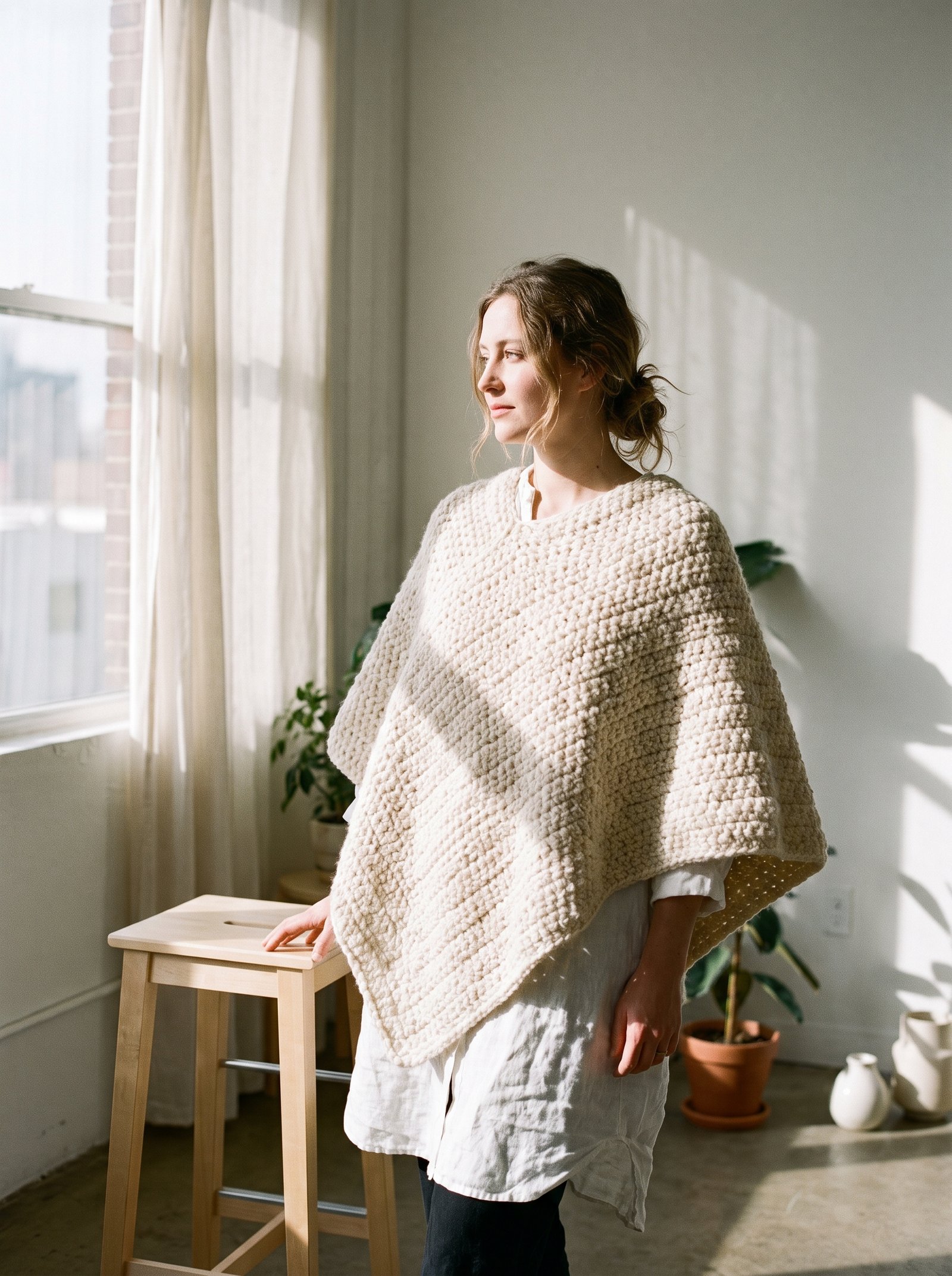 An asymmetrical draped poncho crocheted in undyed natural cream wool, worn effortlessly over a white linen tunic in a bright minimalist studio
