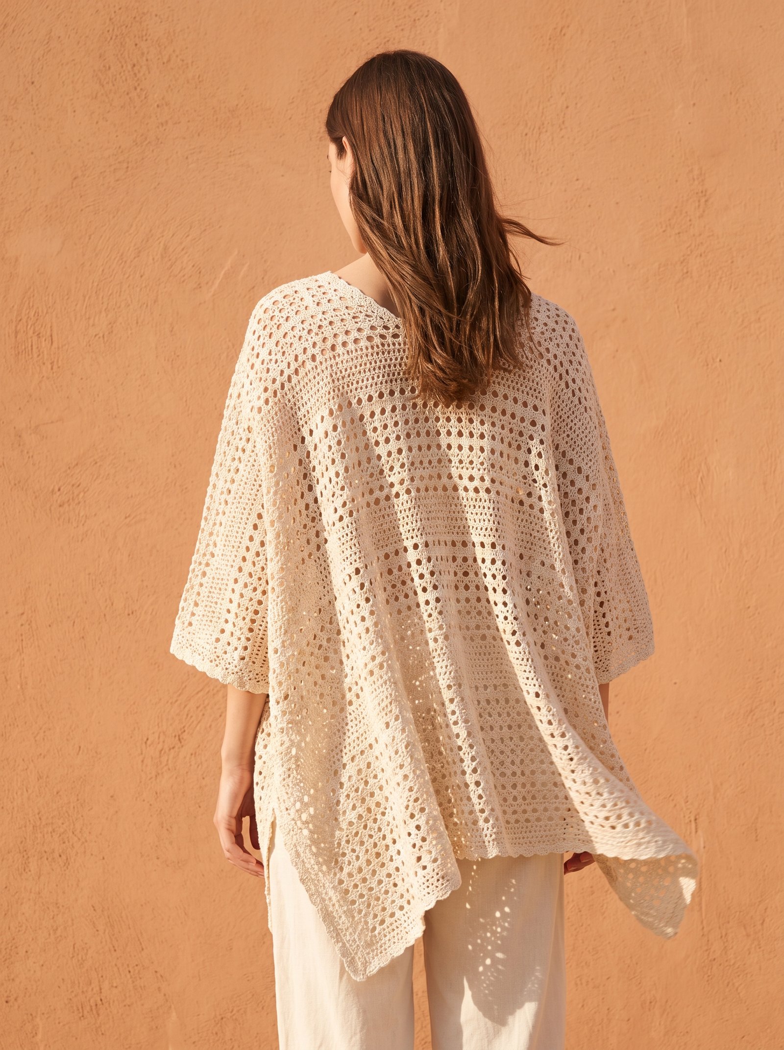 An airy summer lace crochet poncho in unbleached ecru cotton with delicate open eyelet stitches, draped over a model's shoulders against a warm stucco wall