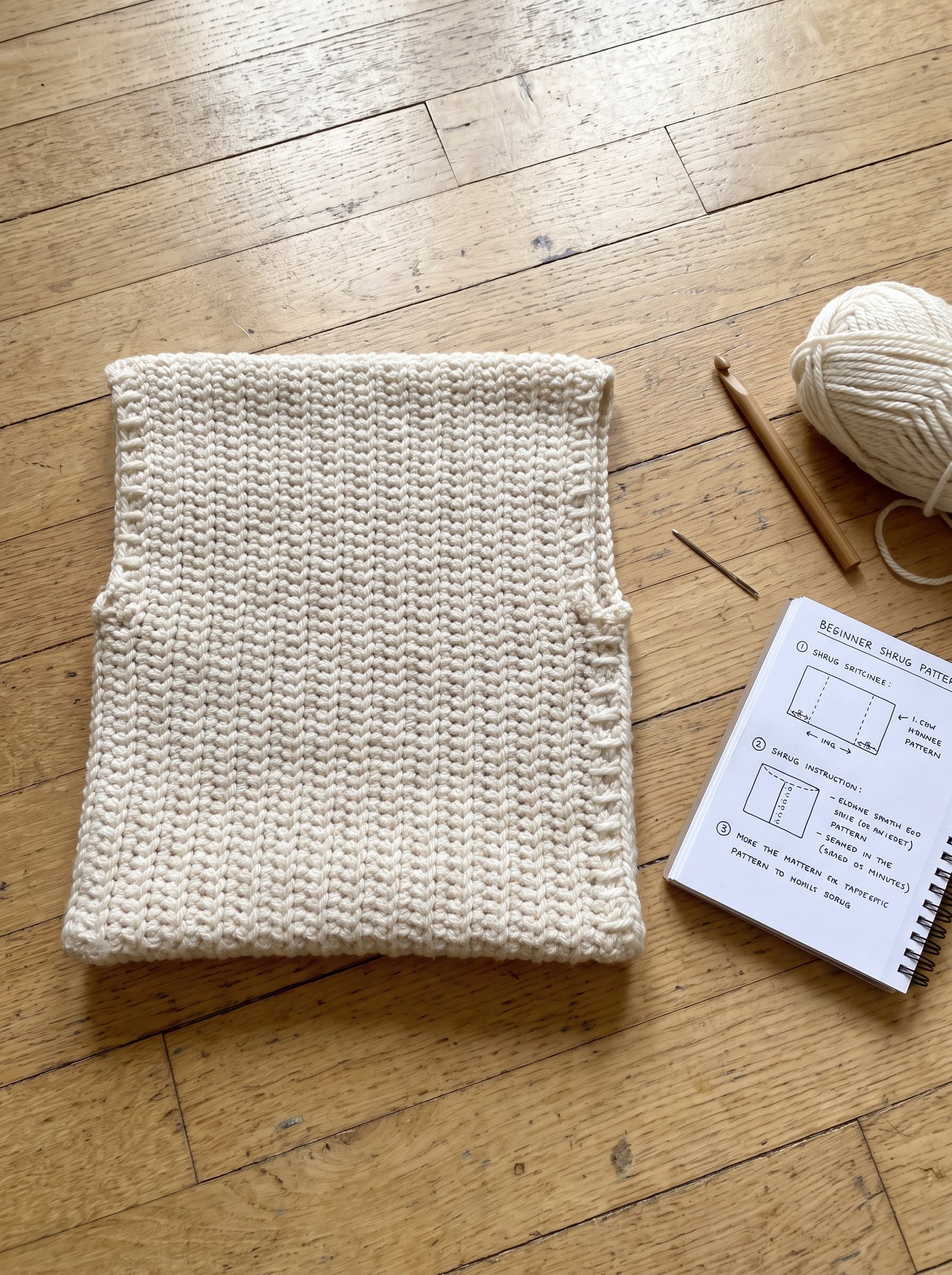Flat lay of a folded textured crochet rectangle showing the side seams creating sleeve openings on a light oak floor