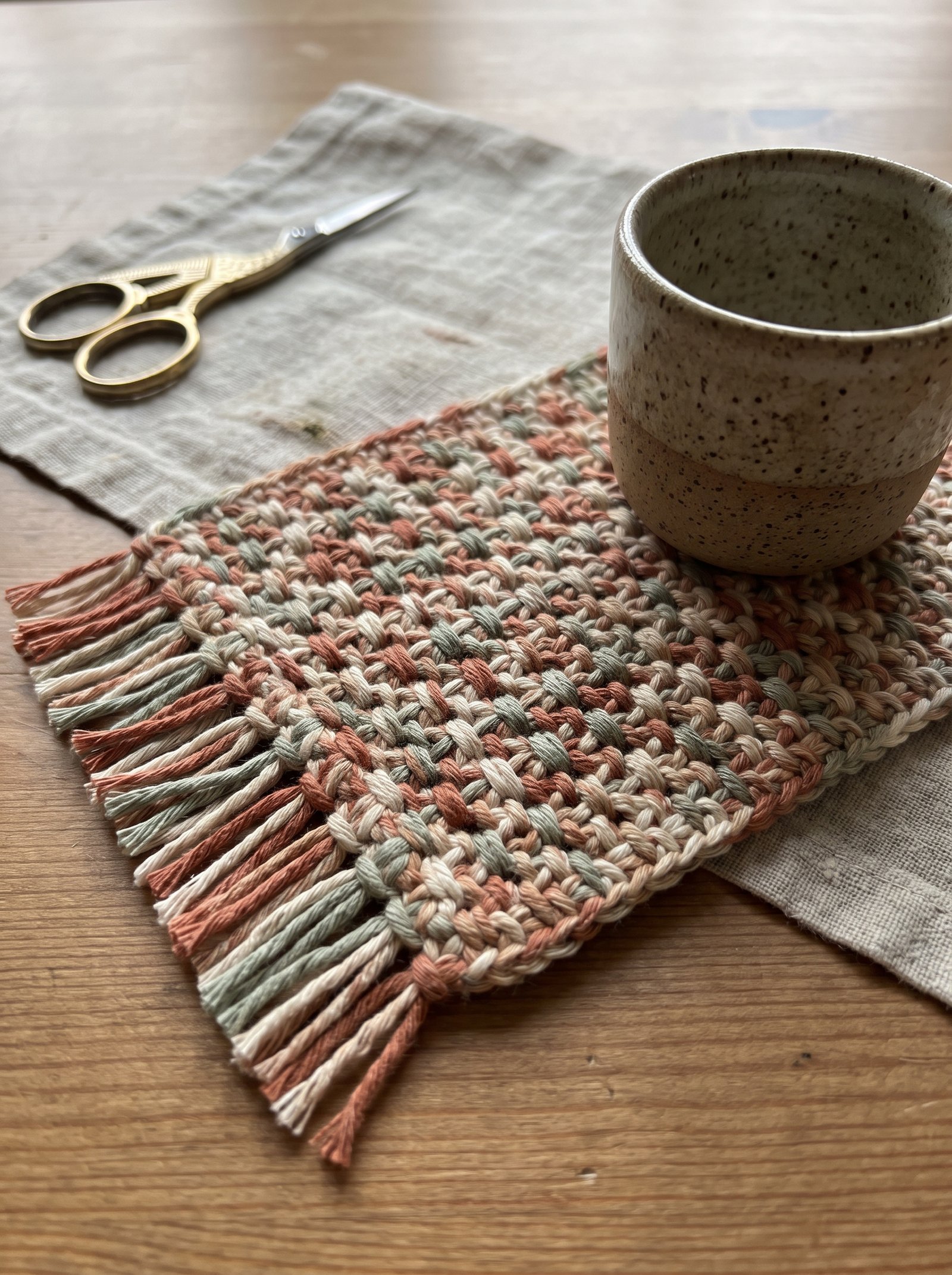 Rectangular moss stitch crochet mug rug with combed fringe beside a rustic ceramic espresso cup