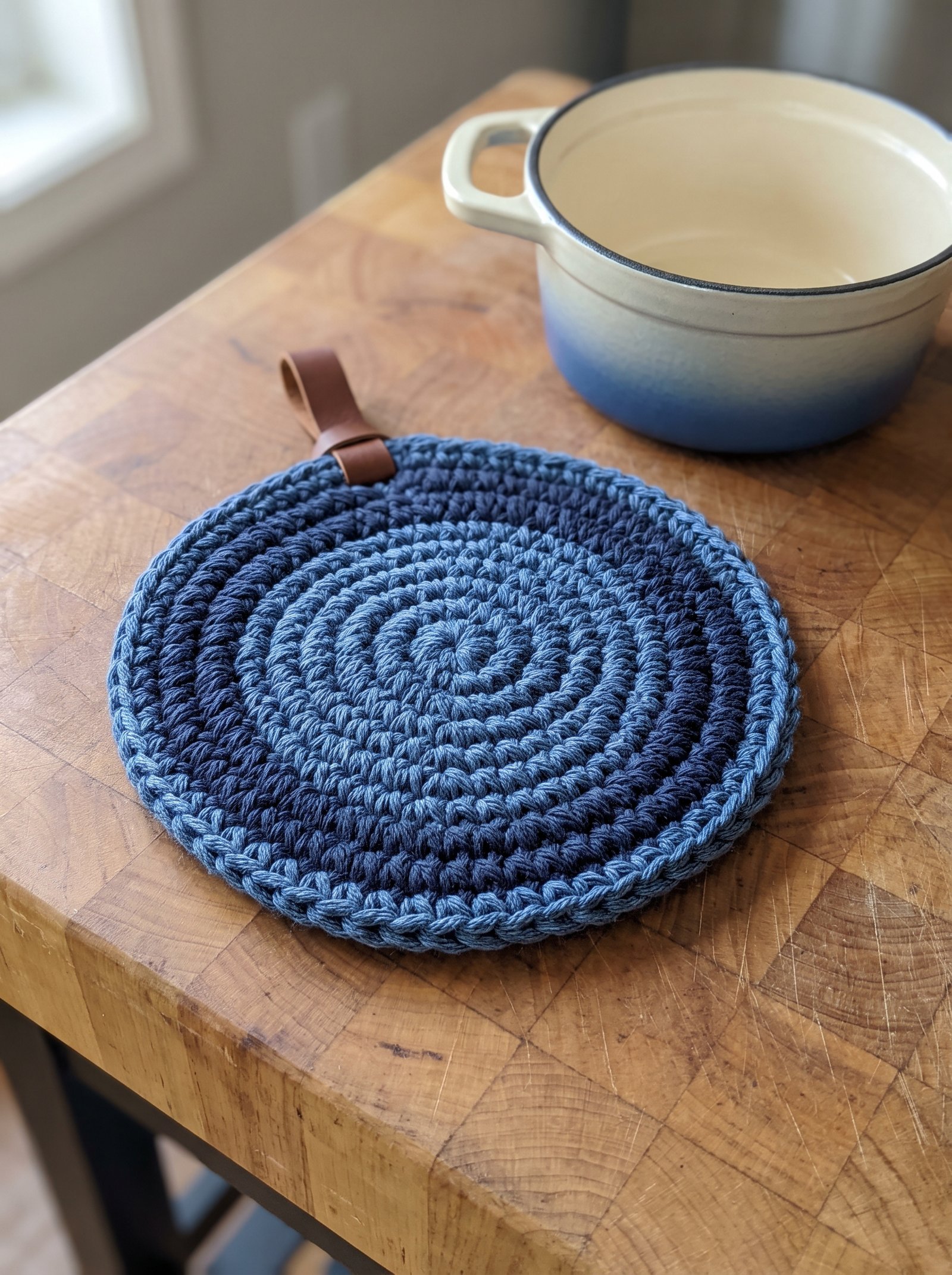 Round navy and denim crochet thermal hot pad trivet with a leather hanging loop on a kitchen island