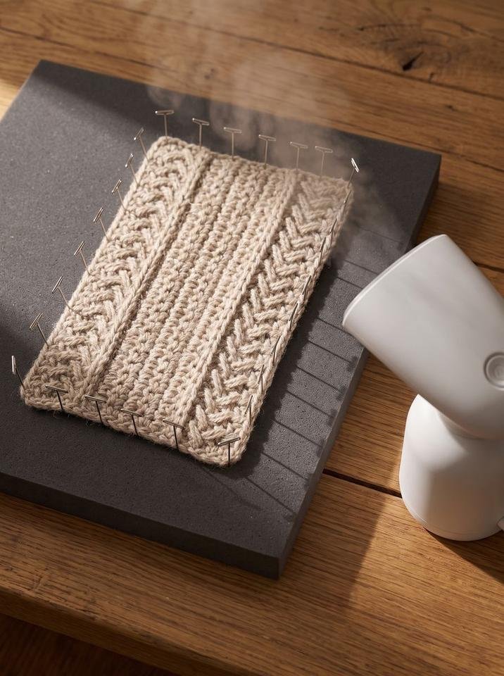 Herringbone crochet swatch pinned to a dark foam blocking mat with steam hovering above it