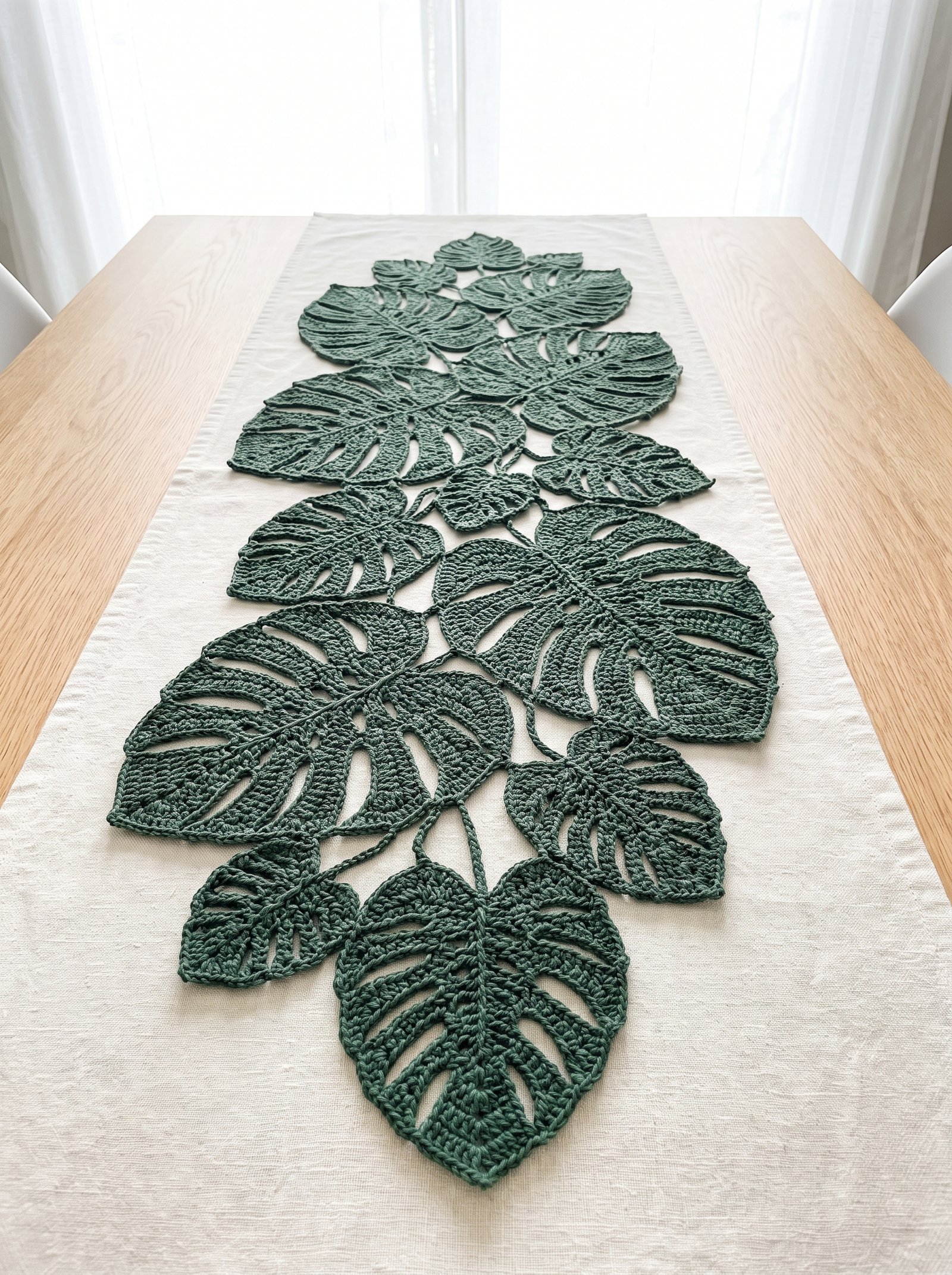 An elaborate dining table runner crafted from interconnected forest green crochet monstera leaves resting on a natural oatmeal linen cloth