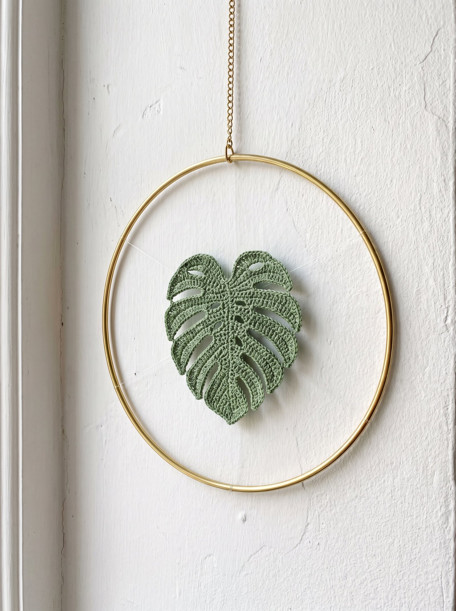 A minimalist sage green crochet monstera leaf suspended inside a polished brass ring by transparent thread hanging on a delicate brass chain
