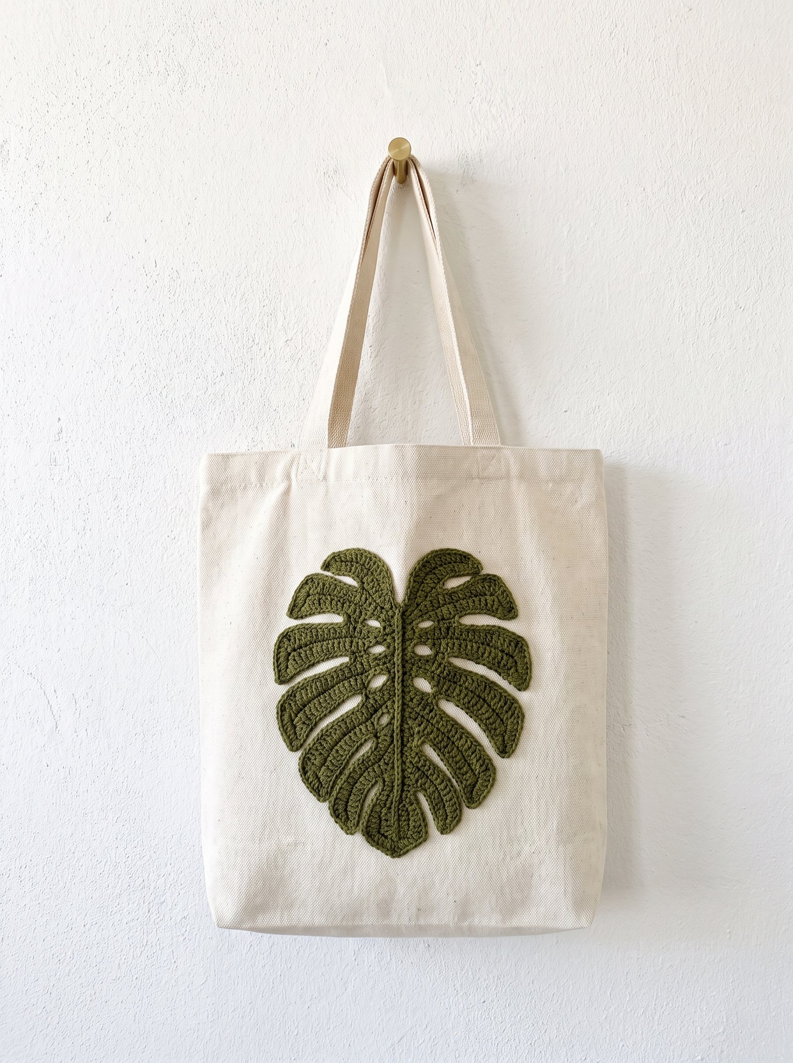 An olive green fenestrated crochet monstera leaf appliqué stitched onto a heavy cream canvas tote bag hanging on a brass coat peg