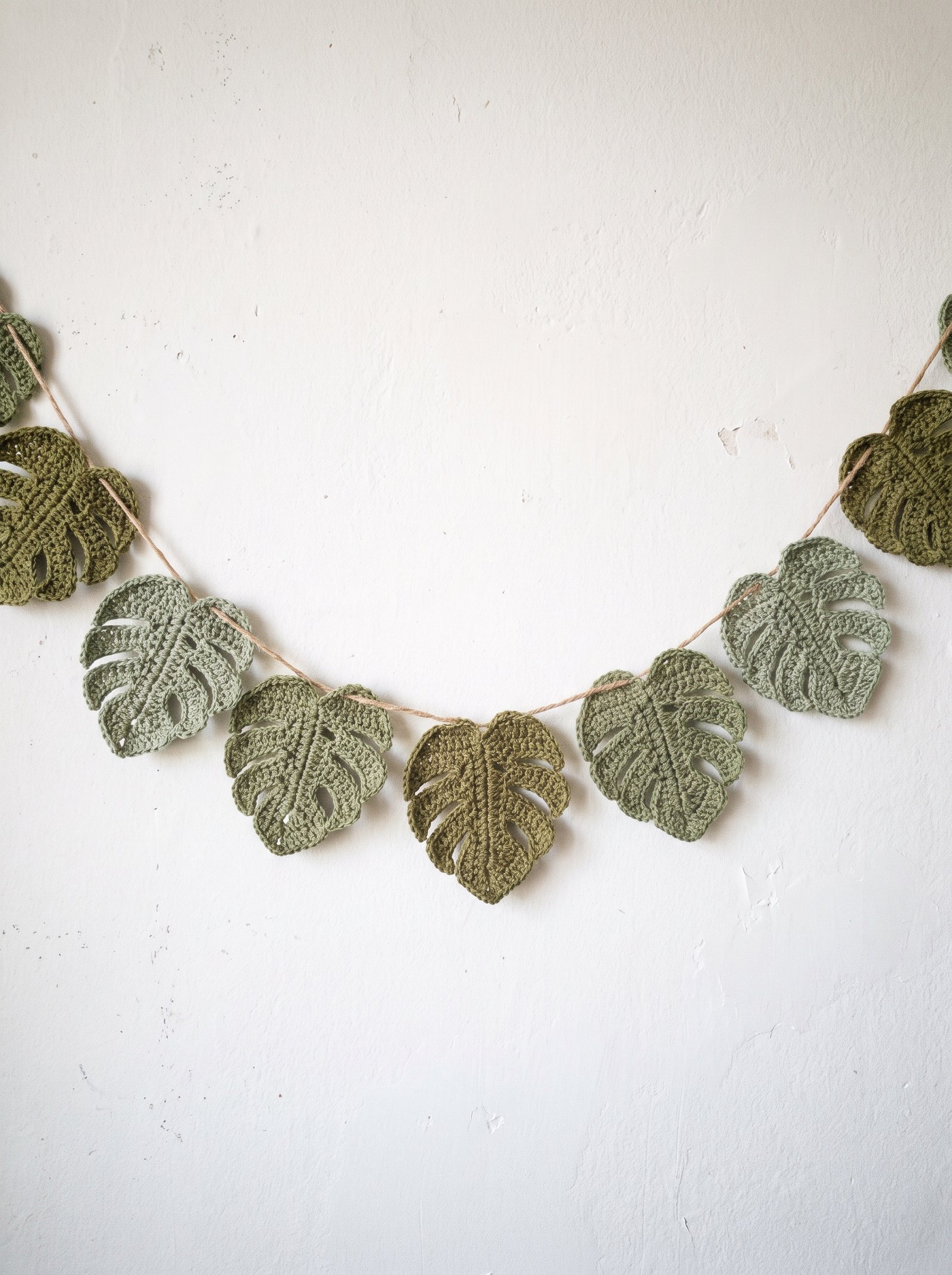 A festive botanical crochet monstera leaf garland strung along rustic jute twine draped gracefully across a bright white plaster wall