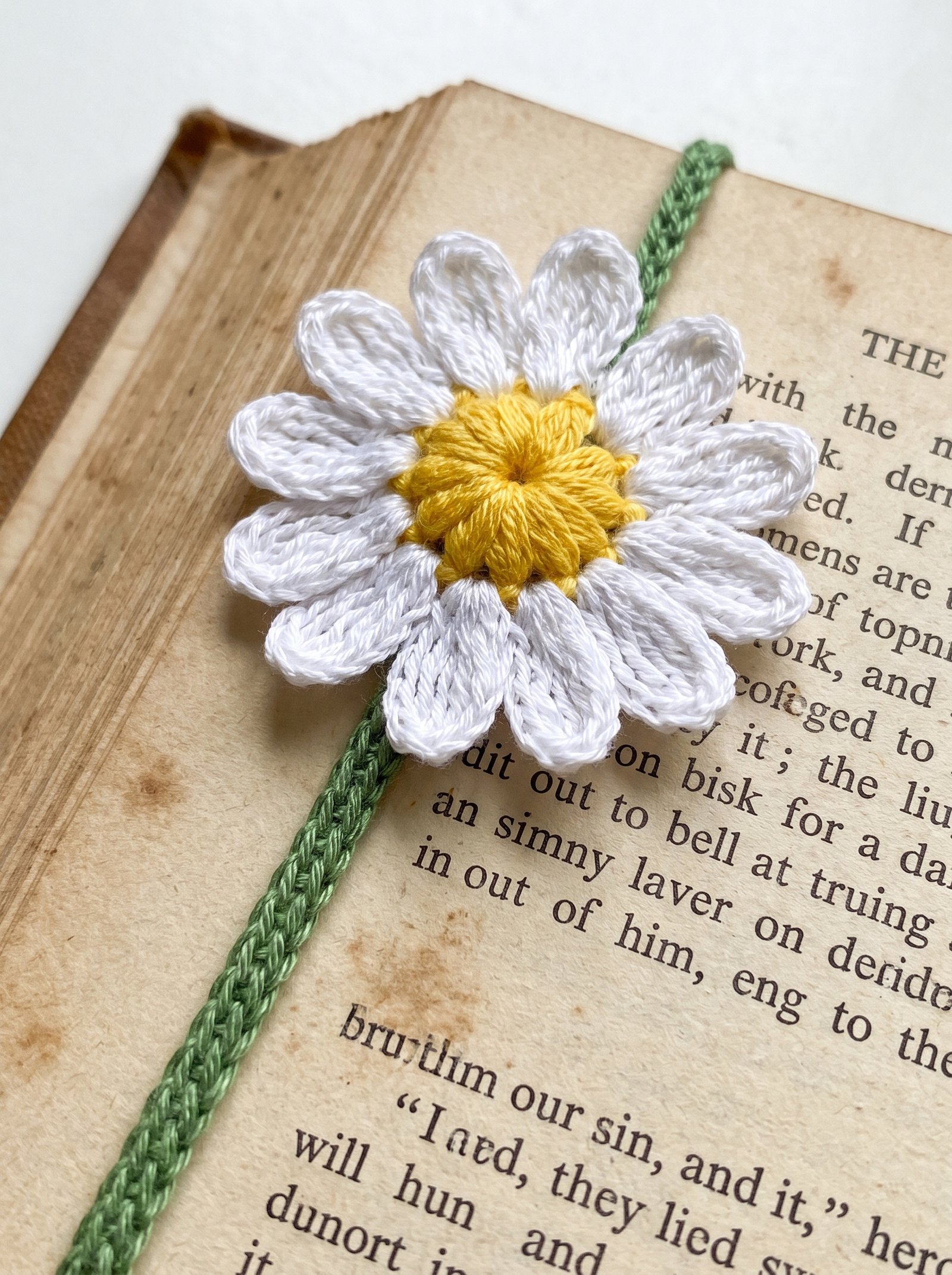 Dainty crochet daisy with white petals and a green chain stem resting inside an open book